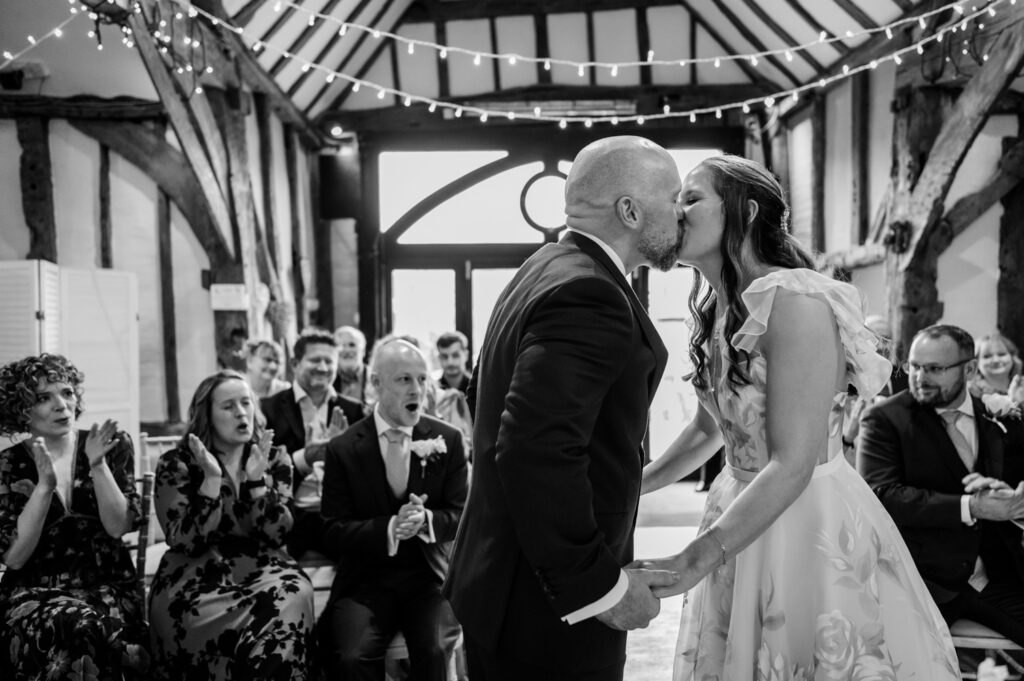 Kristin and Simon sharing their first kiss as a married couple during their intimate Old Luxters Barn micro-wedding elopement, surrounded by smiling guests.